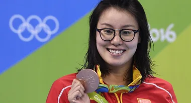 Chinese swimmer smiling, holding a bronze medal at the Rio 2016 Olympics, Olympic rings visible in the background.