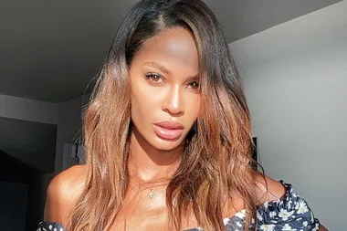 A woman with long brown hair and a floral top, looking at the camera in a sunlit room.