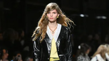 A fashion model walks the runway in a shiny black coat with a white lace collar and long wavy hair.