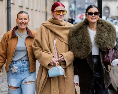 Three women in stylish winter outfits walking on a city street, smiling and carrying handbags.