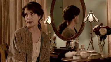 Woman in vintage attire sits by a vanity mirror with flowers, a lamp, and assorted toiletries on the table.