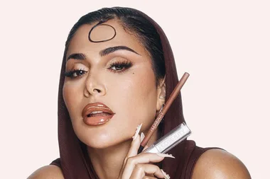 Woman in a brown hood holding Huda Beauty lip products, with glossy lips and styled hair curl on forehead.