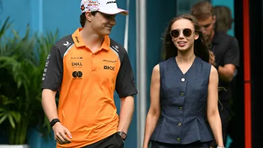 A young man wearing an orange McLaren f1 team polo shirt and baseball hat walking with a young woman wearing round sunglasses and a grey vest.