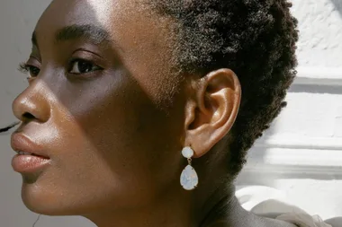 Close-up of a person with short curly hair wearing white gemstone drop earrings, lit by sunlight.
