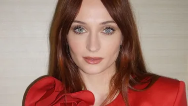 A person with long red hair wearing a red top, posing against a plain background.